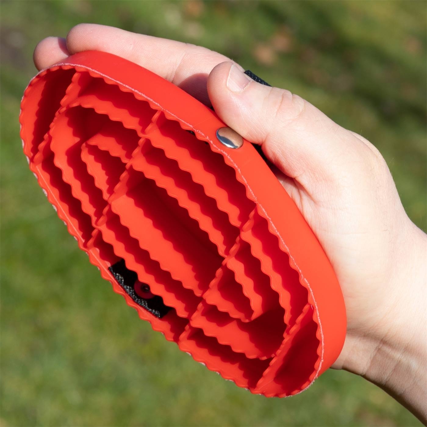 Een hand houdt de HAAS currykam New Generation voor paarden en pony's grooming borstel van Haas van rood rubber met geribbelde haren buiten in een weiland.