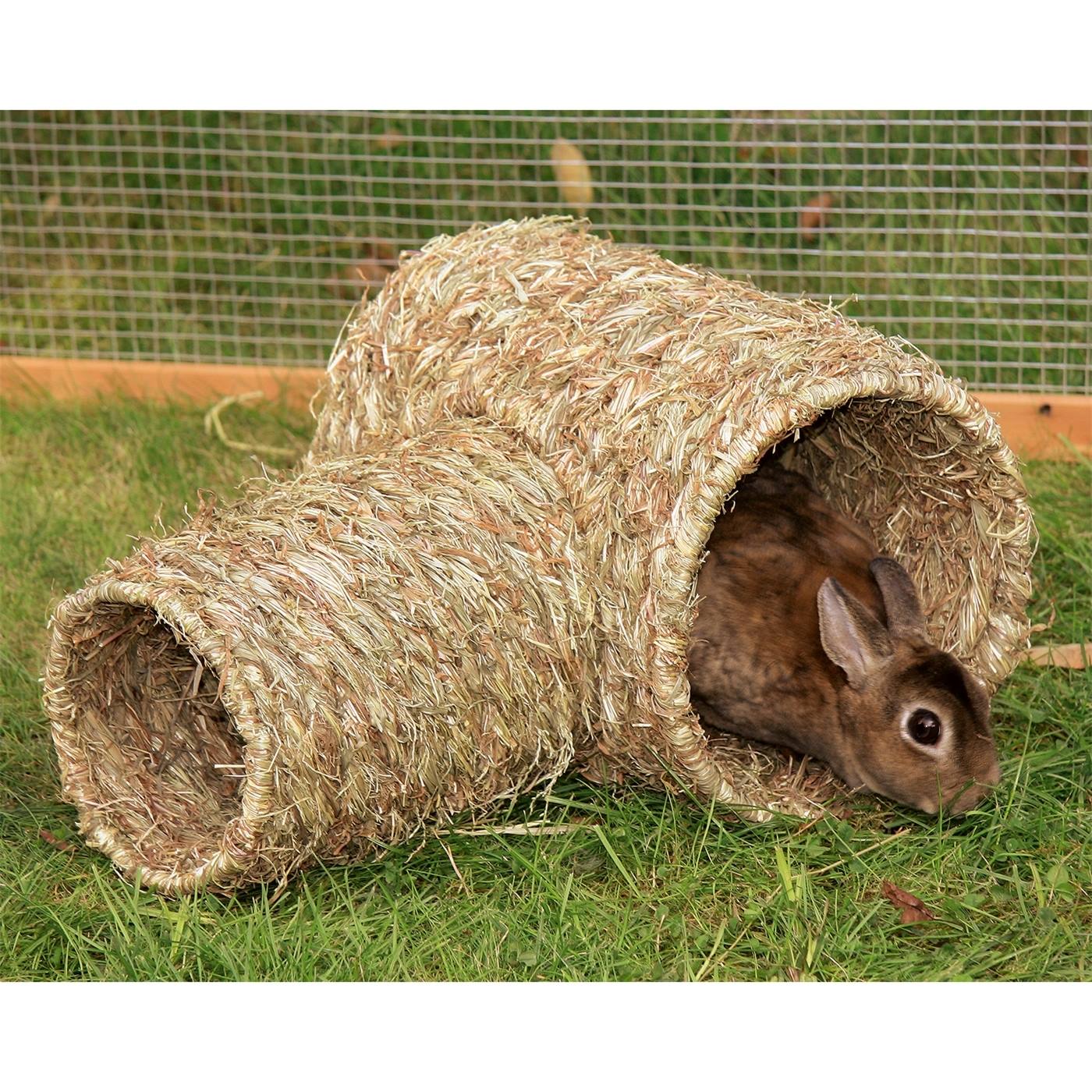 Een bruin konijn zit in de 2-delige graskoker, een geweven grastunnel, op groen gras met een gaashek op de achtergrond.