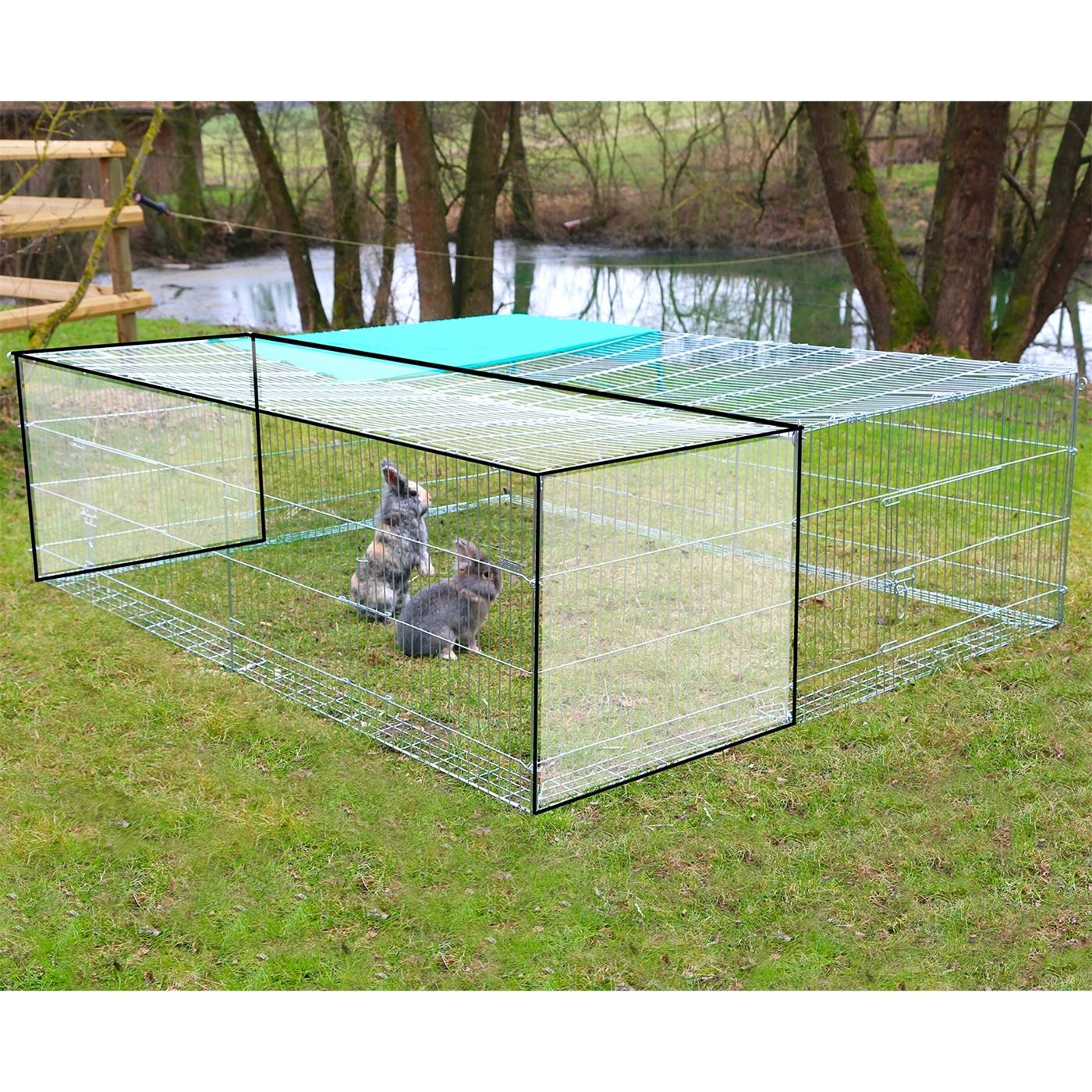 Twee konijnen in een traliehok met het "Uitbreidingselement voor vrije uitloop met ontsnappingsbarrière" op gras bij een vijver en bomen.