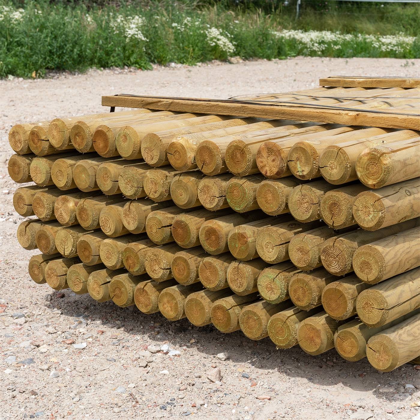 Een pallet ronde houten afrasteringspalen, vanaf achter gefotografeerd en buiten op grind geplaatst, met een achtergrond met bomen en planten.