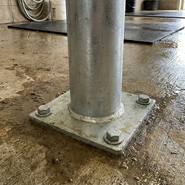 De Growi®-verankeringspaal van 170 cm met ronde buis en voetplaat (Ø 102 mm) wordt met vier grote bouten in een natte betonnen ondergrond verankerd; op de achtergrond zijn water en een slang te zien.